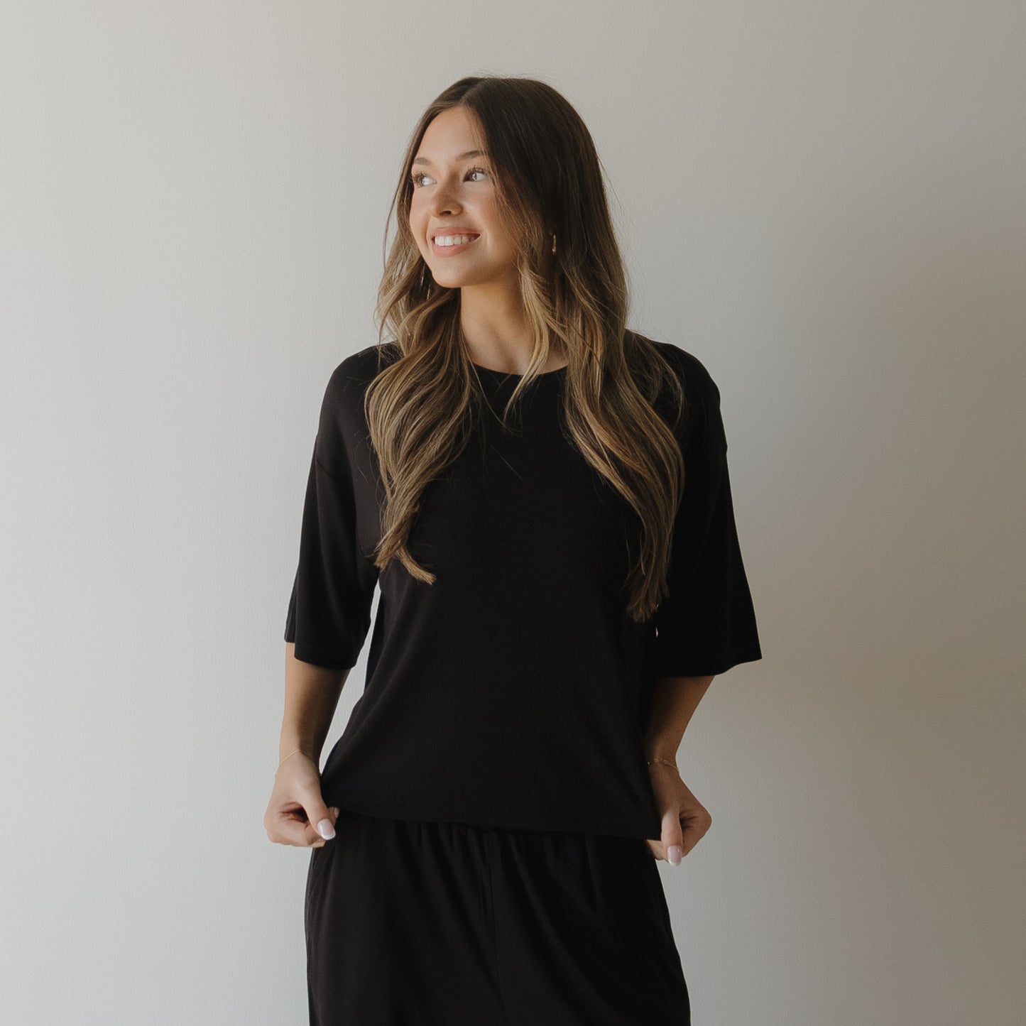 A woman with long, wavy brown hair smiles and looks left while wearing the forever french baby Women's Boxy Tee Lounge Set in ribbed black. She stands against a plain white wall, holding the sides of her top with both hands.