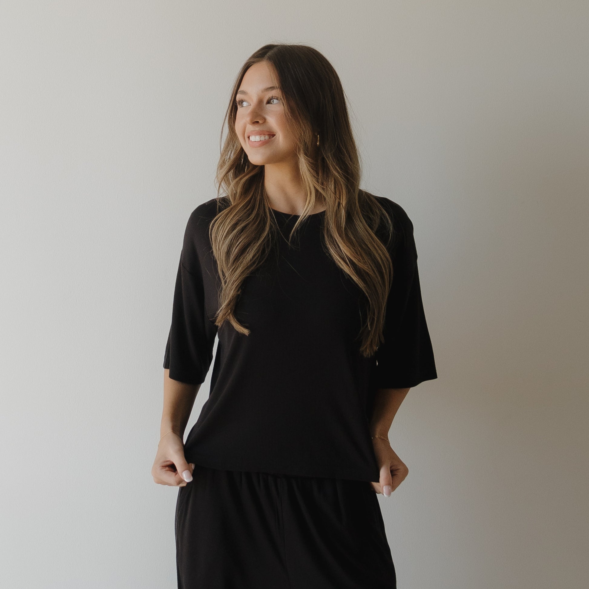 A woman with long, wavy brown hair smiles and looks left while wearing the forever french baby Women's Boxy Tee Lounge Set in ribbed black. She stands against a plain white wall, holding the sides of her top with both hands.