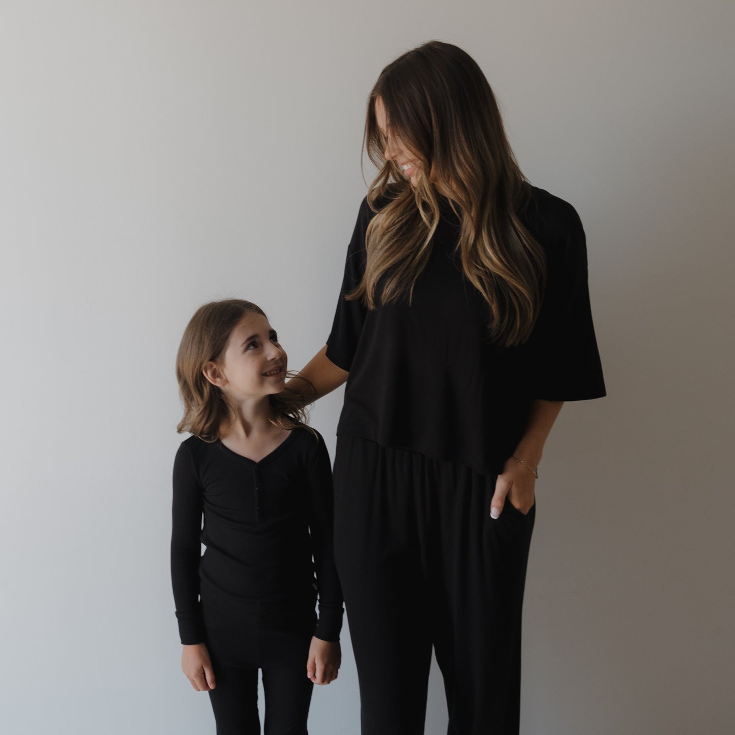 A woman and a young girl with long brown hair smile at each other, both wearing forever french baby Bamboo Two Piece Pajamas | Ribbed Black, standing against a plain light-colored wall.