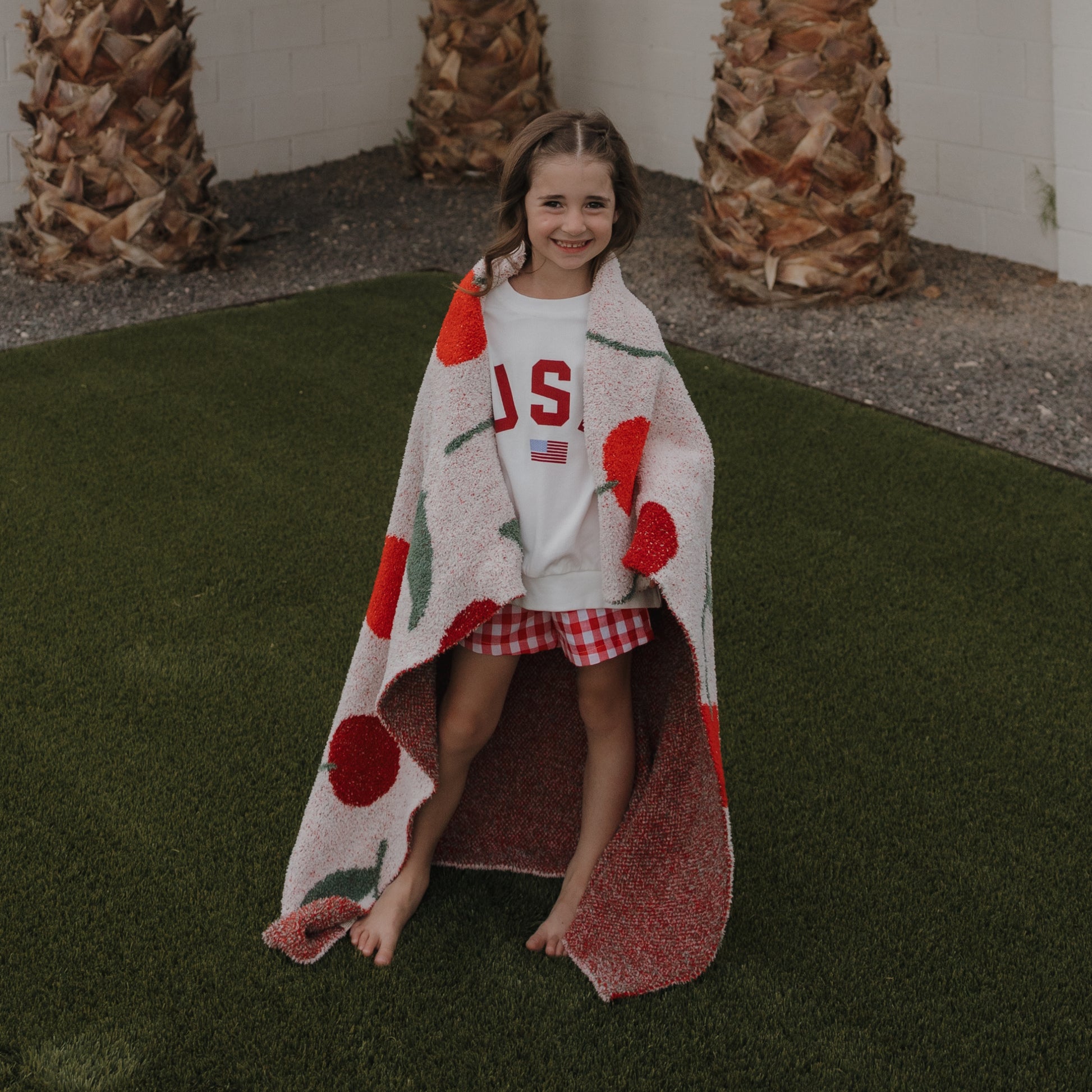 A smiling girl stands on artificial grass, wrapped in the forever french baby Plush Blanket | Cherry. She wears a white “USA” T-shirt and red checkered shorts, with palm tree trunks and a white wall in the background.