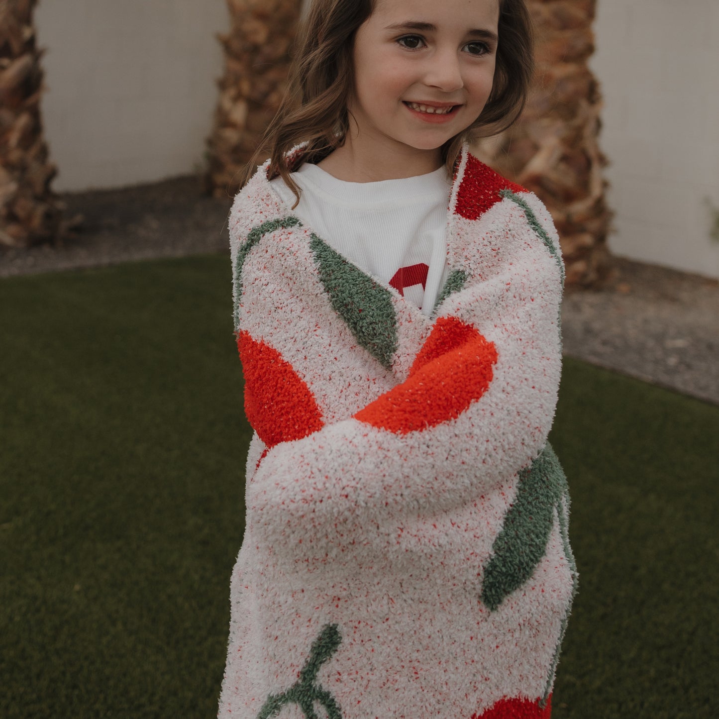A young child smiles outdoors, wrapped in the forever french baby Plush Blanket | Cherry, featuring red and green cherry patterns. Palm trees and a grassy lawn are visible in the background.