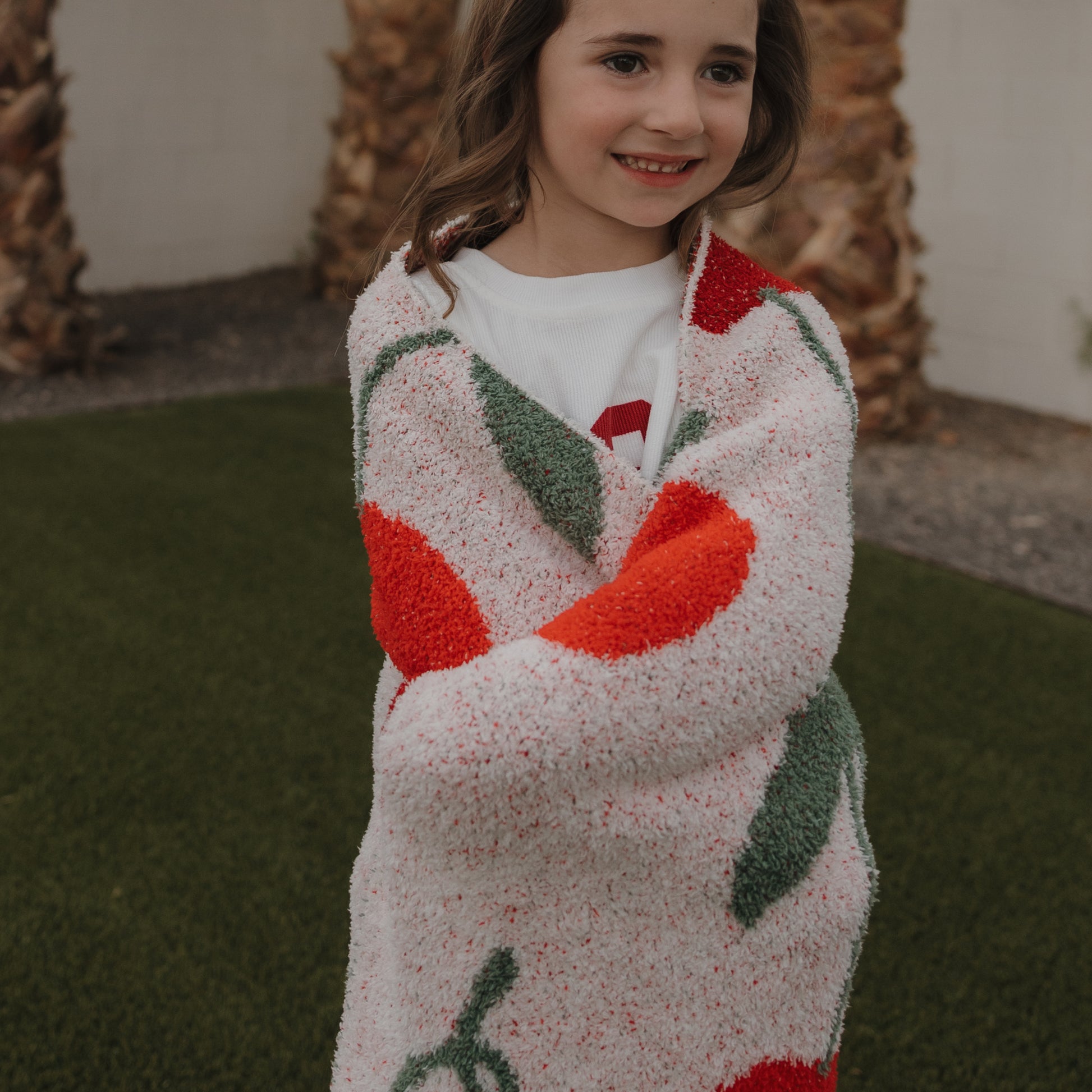 A young child smiles outdoors, wrapped in the forever french baby Plush Blanket | Cherry, featuring red and green cherry patterns. Palm trees and a grassy lawn are visible in the background.