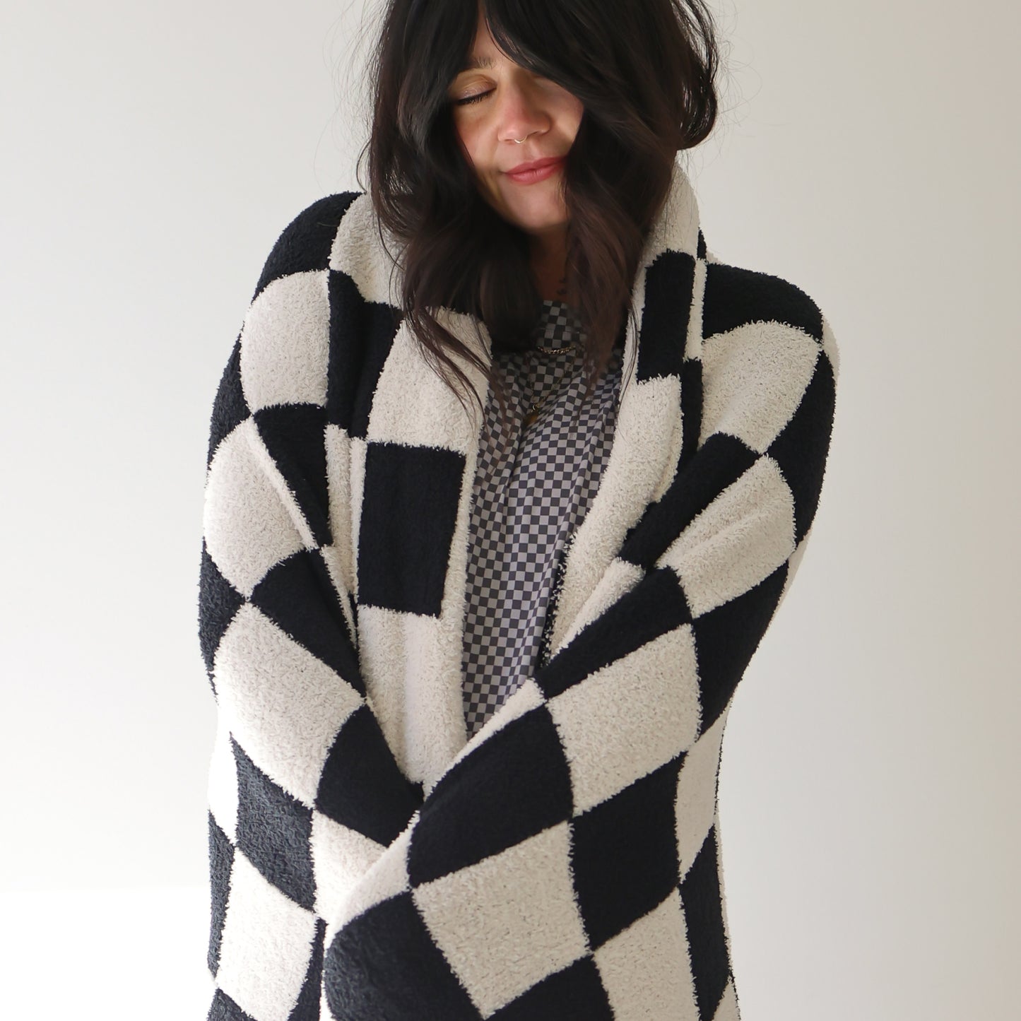 A woman with dark hair stands against a light background, cozy and content, wrapped in the forever french baby Plush Blanket in Black & White Checkerboard.