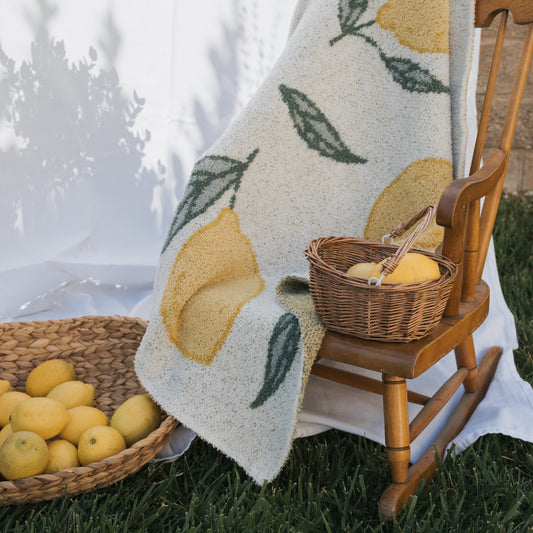 A *PRE-ORDER* Plush Blanket | Lemon by forever french baby, Oeko-tex certified with a lemon and leaf print, is draped over a wooden chair beside wicker baskets of lemons, all set on grass with a white fabric backdrop.