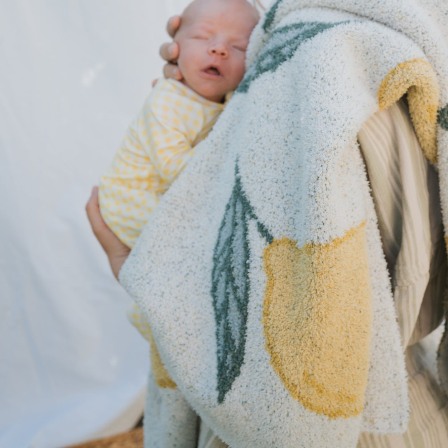 A newborn in a yellow polka-dot onesie is cradled by an adult wrapped in the forever french baby *PRE-ORDER* Plush Blanket | Lemon, featuring Oeko-Tex certified softness and yellow-green shapes, against a light background.