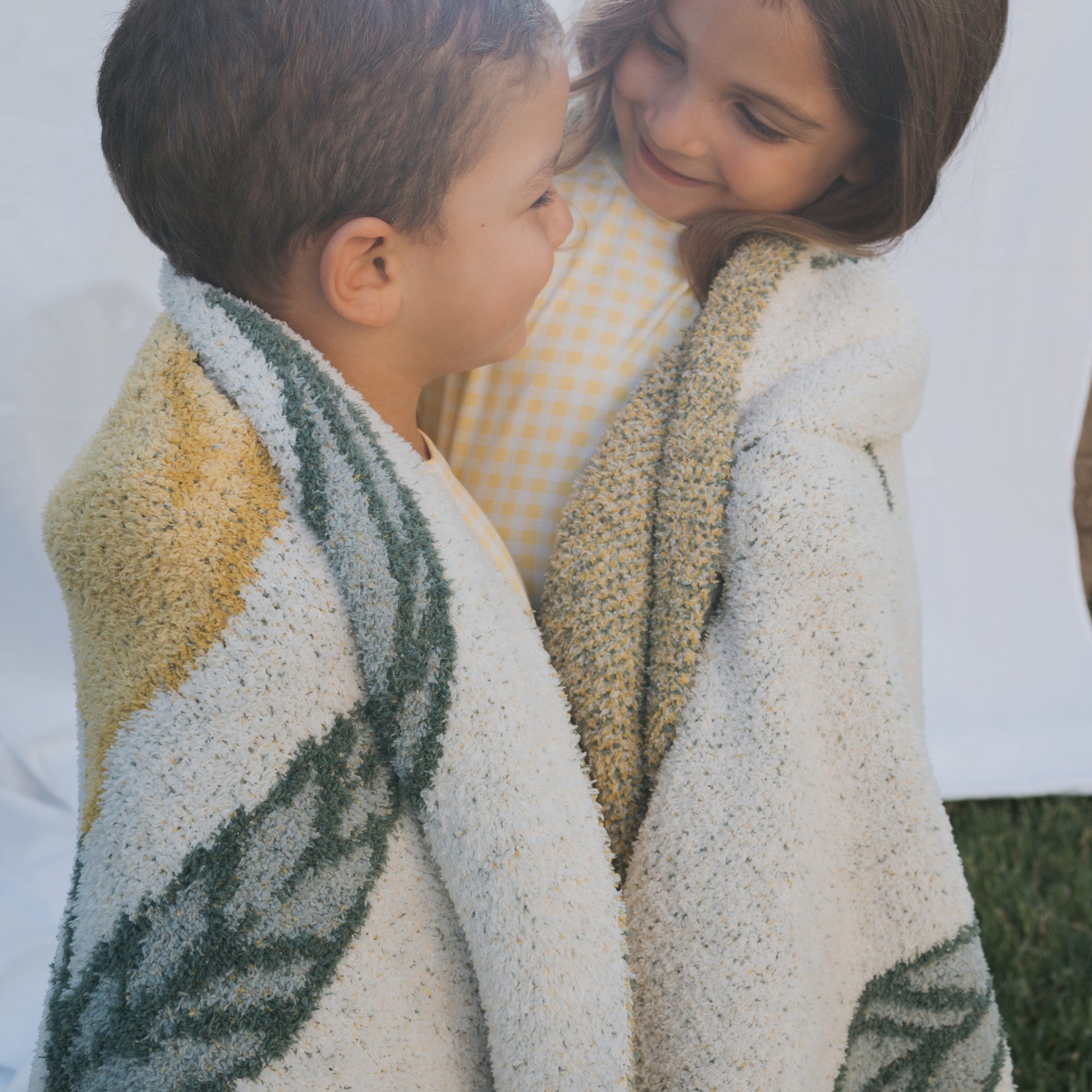 Two smiling children, a boy and a girl, snuggle together in the forever french baby *PRE-ORDER* Plush Blanket | Lemon, featuring soft Oeko-tex certified fabric with cheerful green and yellow patterns.