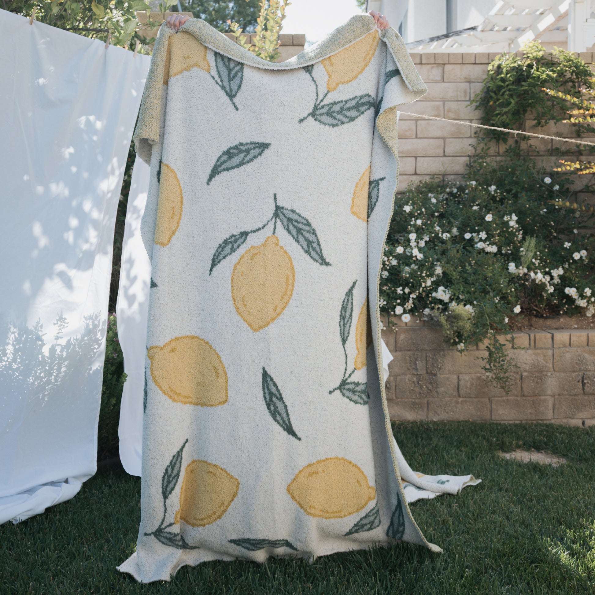 A person holds up the forever french baby *PRE-ORDER* Plush Blanket | Lemon, featuring yellow lemons and green leaves, while standing on grass in a garden with a brick wall, white flowers, and white sheets behind them.