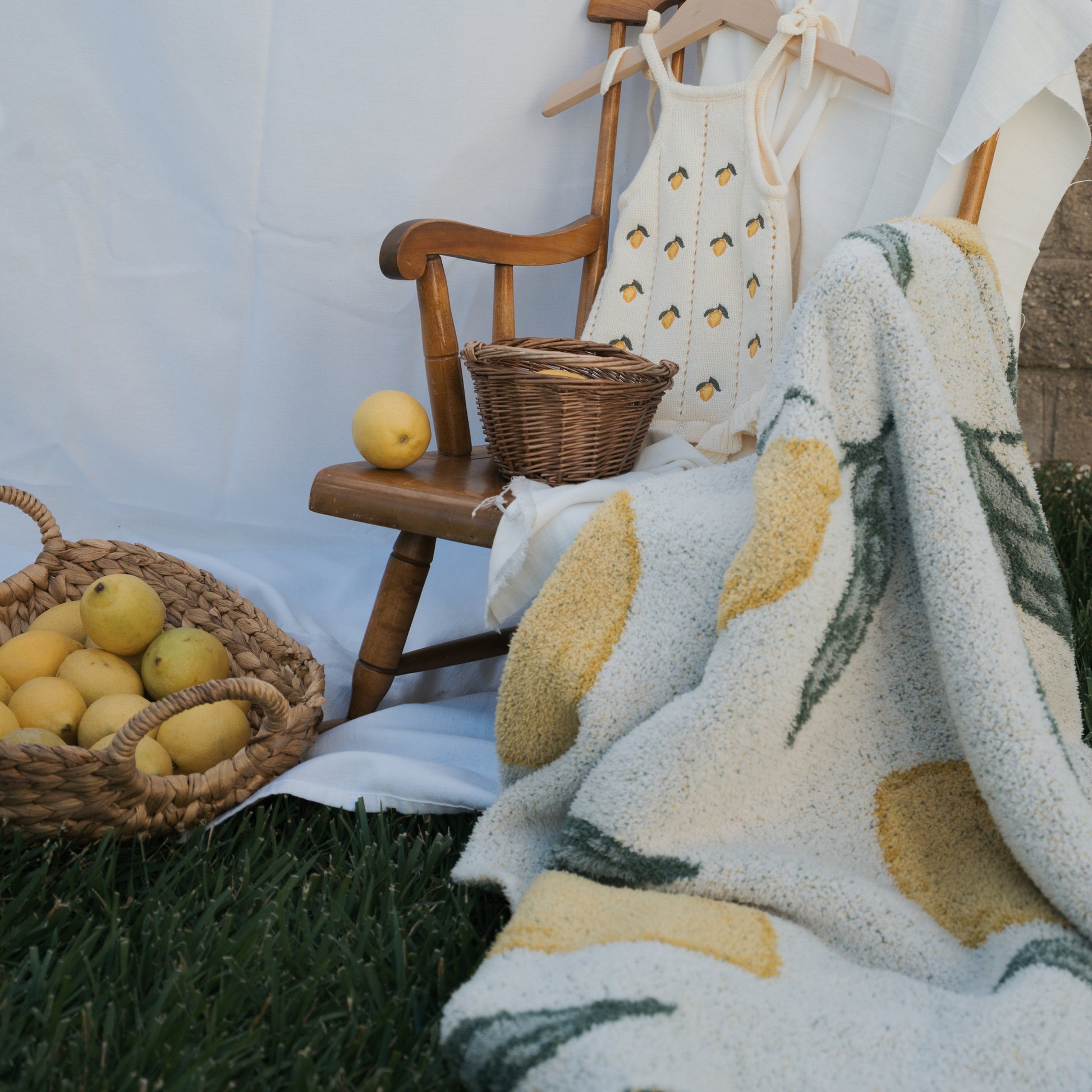 A wooden chair with a lemon-patterned dress and wicker basket sits on grass beside a woven basket of lemons and the forever french baby *PRE-ORDER* Plush Blanket | Lemon, Oeko-tex certified, all set against a white backdrop.