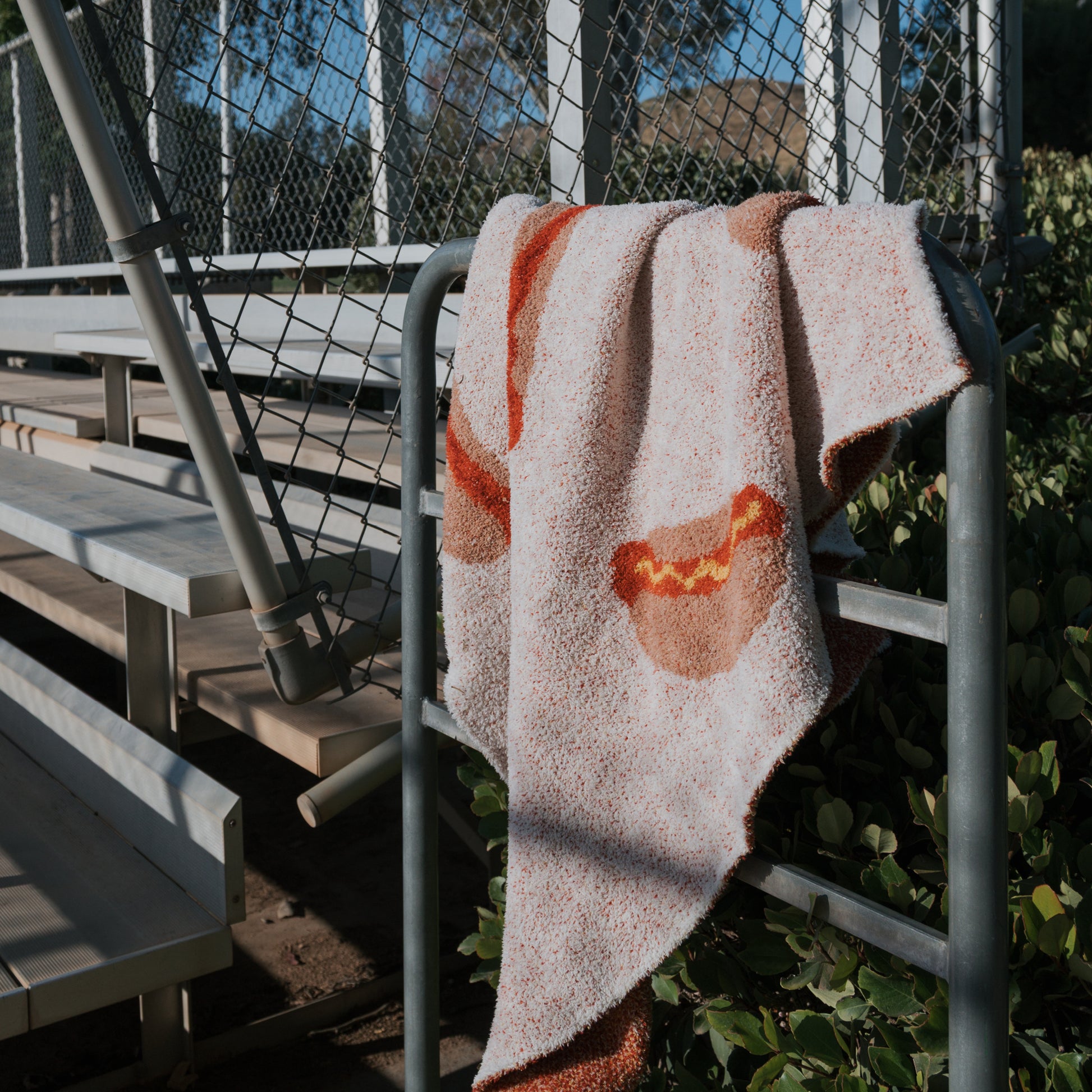 The forever french baby Plush Blanket | Hot Dogs, featuring playful hot dog designs, is draped over a metal railing near empty bleachers and a chain-link fence, with greenery and sunlight in the background.