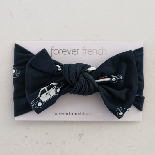 The Bamboo Head Wrap | Police Cruiser by forever french baby is a navy blue bamboo baby headband with white police car patterns, displayed on a branded card.
