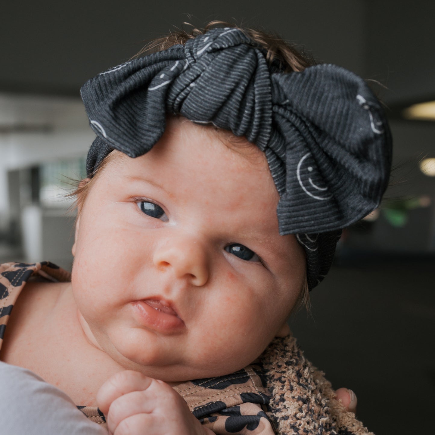 A light-skinned baby with chubby cheeks wears the forever french baby Bamboo Head Wrap | Smiley Ribbed, a large dark gray adjustable bow headband with white smiley faces, looking calmly at the camera.