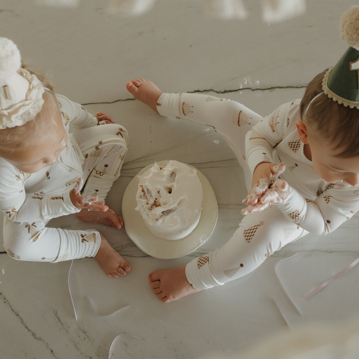 Two toddlers in matching forever french baby Child Sleepy Time Set | Birthday Boy pajamas and party hats sit on the floor around a messy cake, with one child’s hands covered in frosting. A marble floor and soft lighting create a cozy atmosphere.