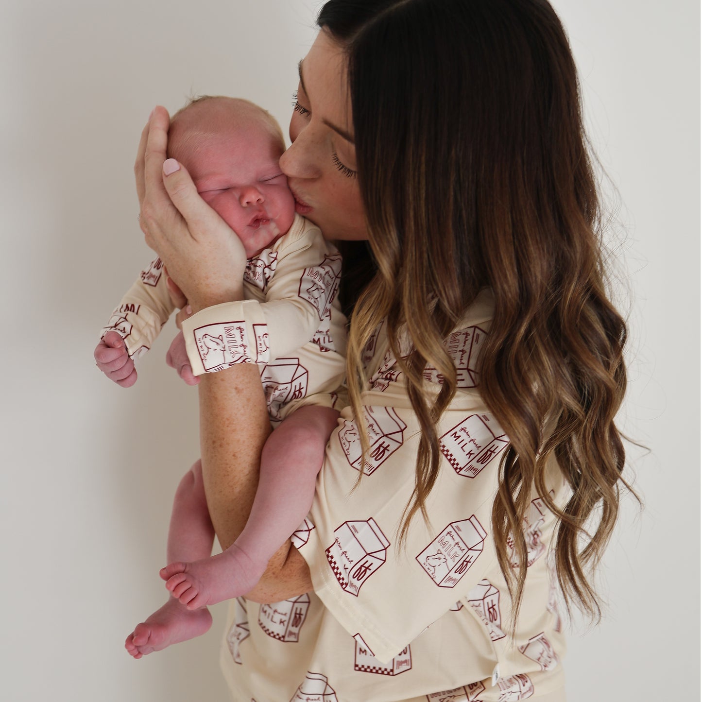 A woman with long brown hair kisses a newborn dressed in the "Long Sleeve Bamboo Snapsuit | Milk" by forever french baby. Both wear matching hypoallergenic outfits against a plain white background.