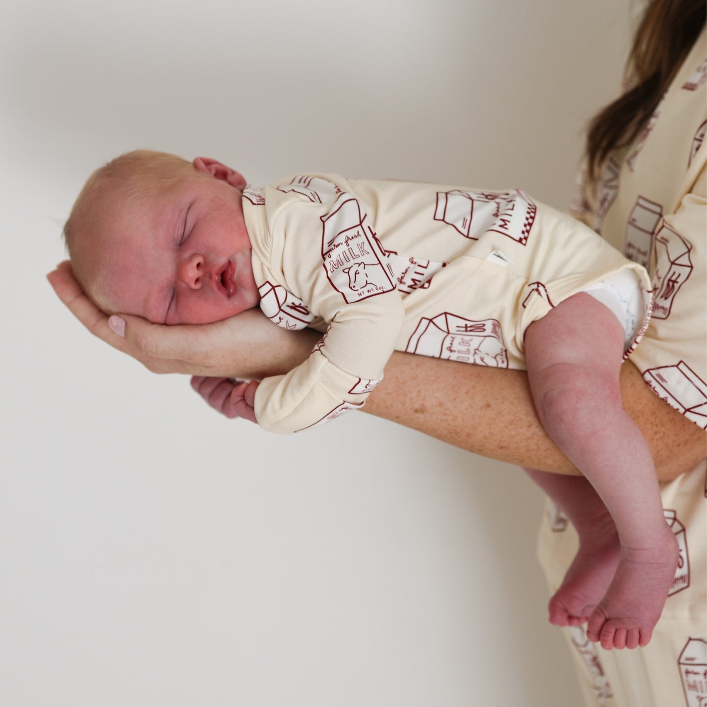 A newborn in a forever french baby Long Sleeve Bamboo Snapsuit | Milk sleeps, cradled gently in an adult's hand and arm, against a plain white background.