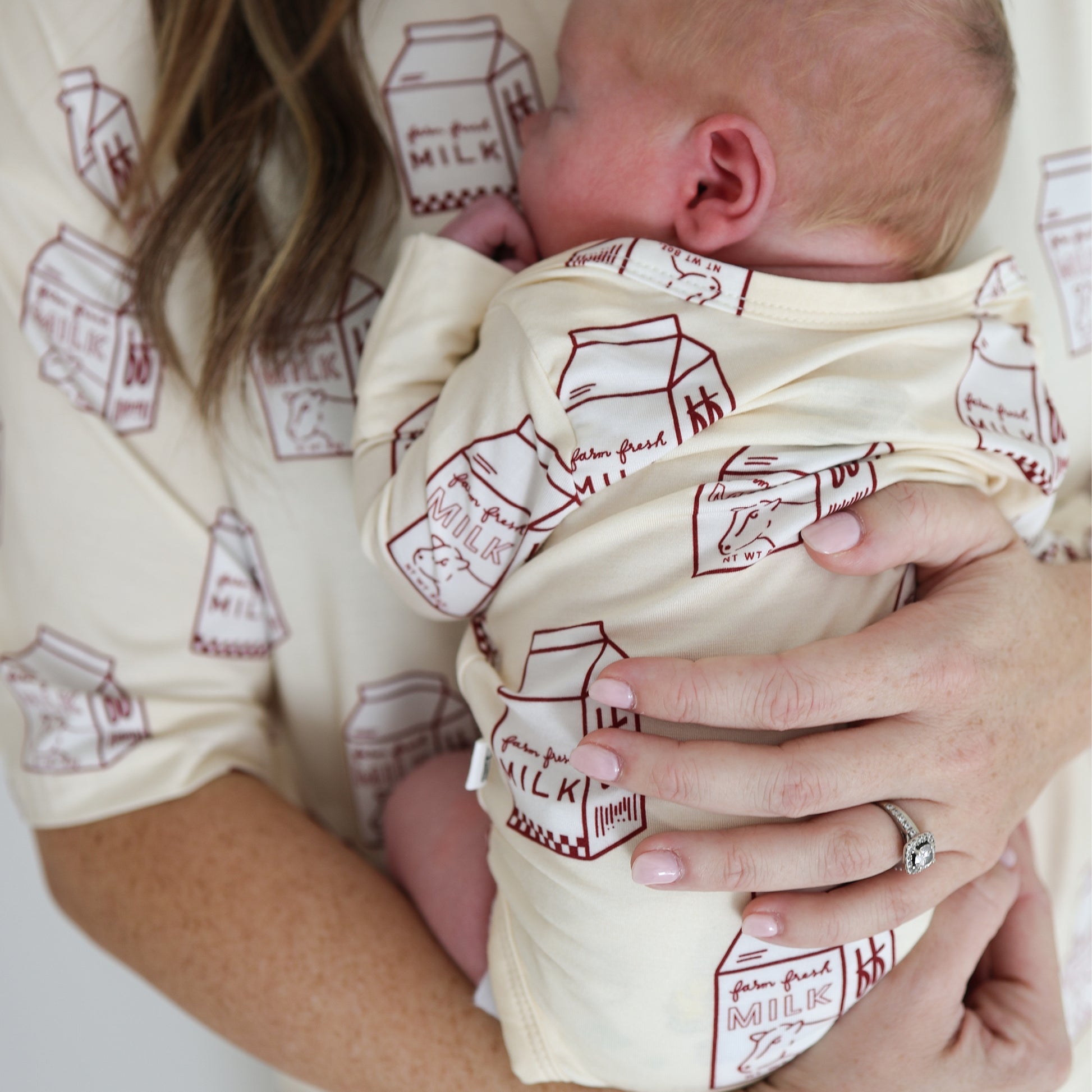 A woman wearing a diamond ring gently supports a sleeping baby dressed in the forever french baby Long Sleeve Bamboo Snapsuit | Milk, featuring matching cream fabric with brown milk carton prints.