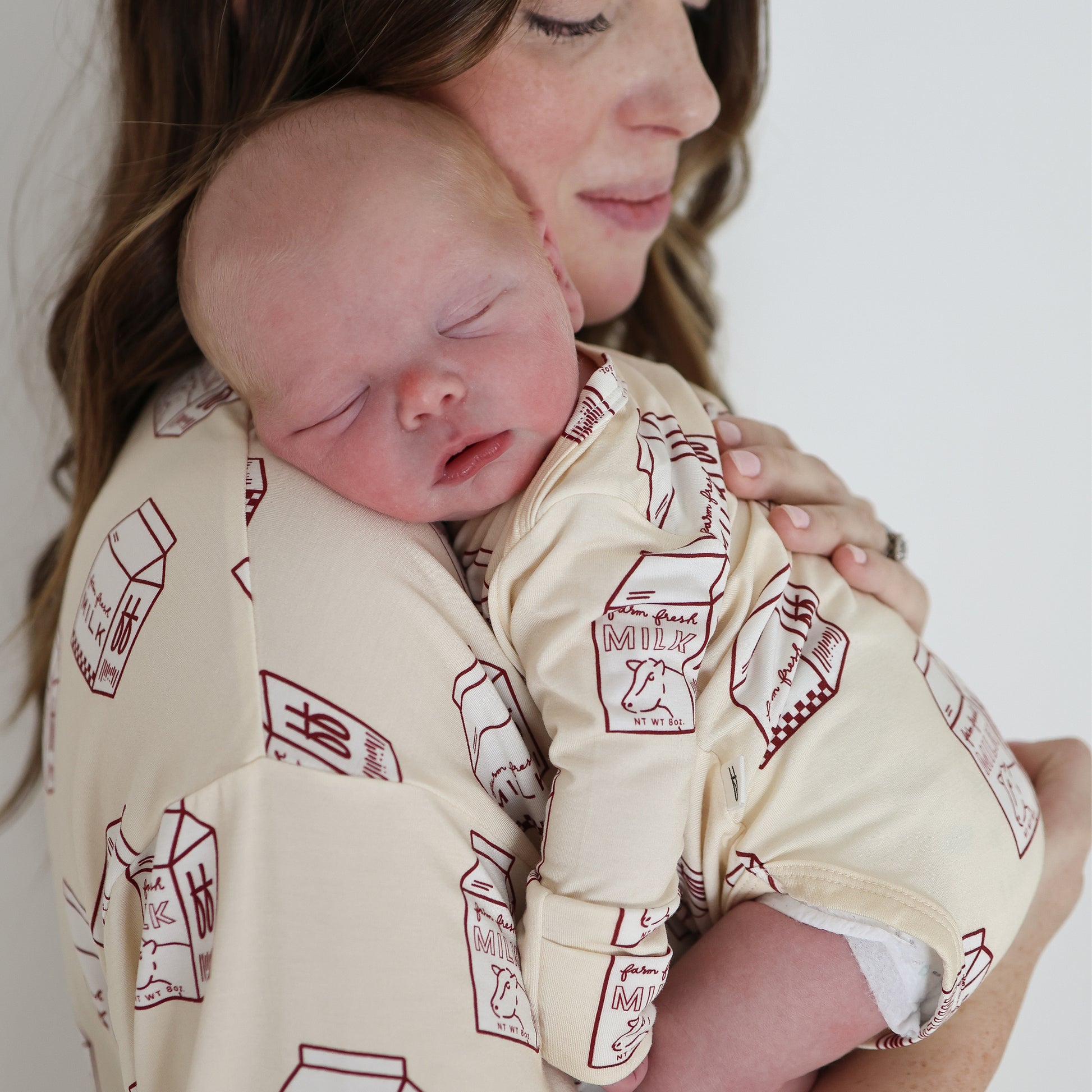 A woman holds a sleeping baby on her shoulder. Both wear matching forever french baby Long Sleeve Bamboo Snapsuits in cream with red milk carton prints. The clothing is hypoallergenic, and the baby's resting peacefully while the woman's face is partly visible.