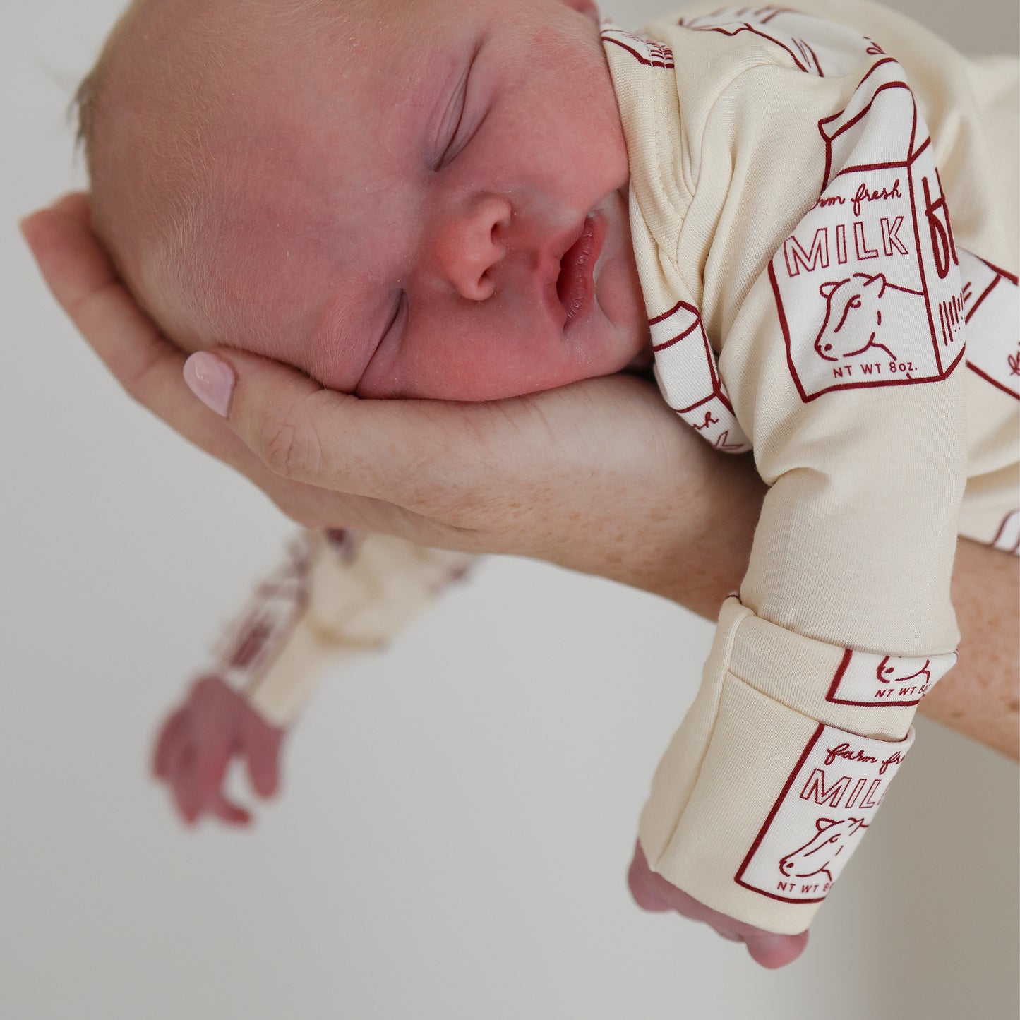 A newborn in the forever french baby Long Sleeve Bamboo Snapsuit | Milk, featuring red milk carton designs, sleeps peacefully cradled in an adult’s hand—ideal for those seeking hypoallergenic baby clothing.