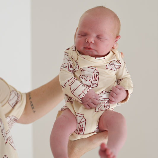 A newborn in the forever french baby Long Sleeve Bamboo Snapsuit | Milk, featuring a milk carton print, is held upright by an adult. The baby's eyes are closed against a plain, light background.