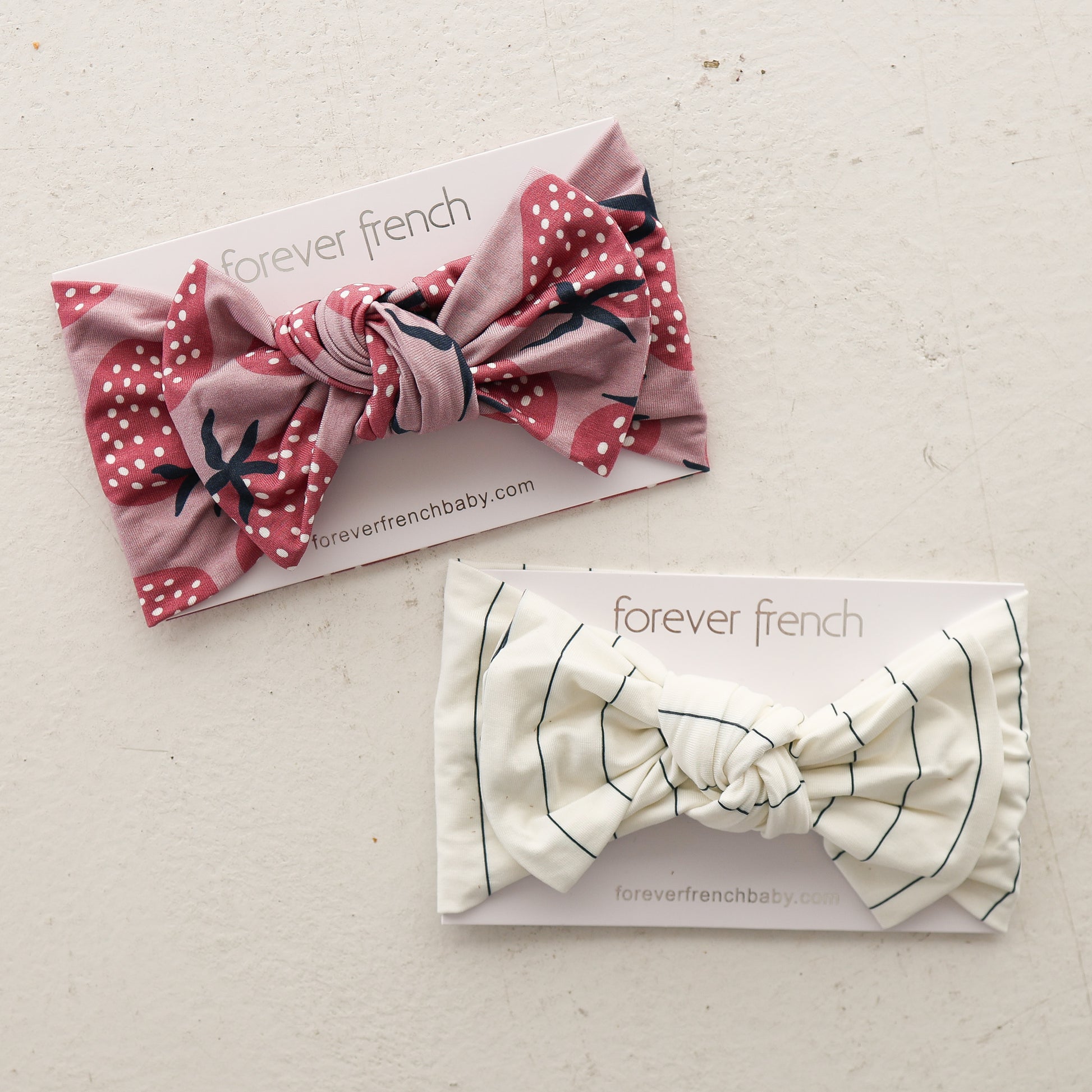 Two Bamboo Head Wrap | Matilda headbands by forever french baby are shown on branded cards: one is pink with white polka dots and florals, the other cream with thin black stripes, all on a light, textured surface.