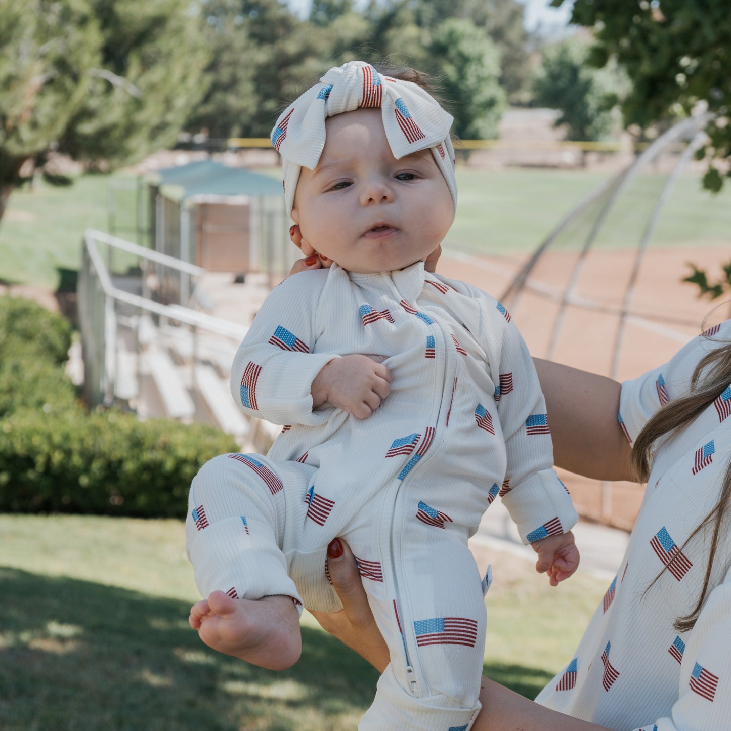 A baby wearing forever french baby’s Bamboo Zip Pajamas | Americana Ribbed—white with American flag prints—is held outdoors near a baseball field with trees and greenery in the background.