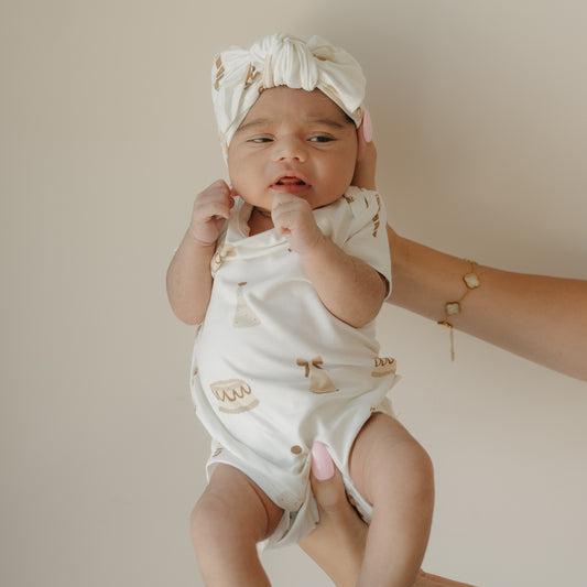 A baby wearing the forever french baby Short Sleeve Bamboo Snapsuit | Birthday Girl—a white, patterned bamboo viscose romper—paired with a matching headwrap is held up by an adult with pink nails and a gold bracelet against a plain light background. This hypoallergenic snapsuit provides exceptional comfort and softness for delicate skin.
