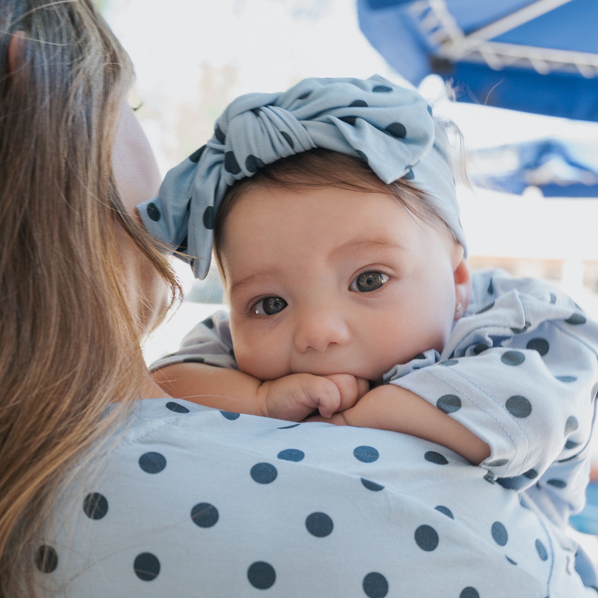 A baby wearing the forever french baby Bamboo Head Wrap in Sully and a matching outfit rests on an adult's shoulder, who is also dressed in a polka-dot top. Gazing at the camera while sucking on their fingers, the baby showcases forever french baby's signature style.
