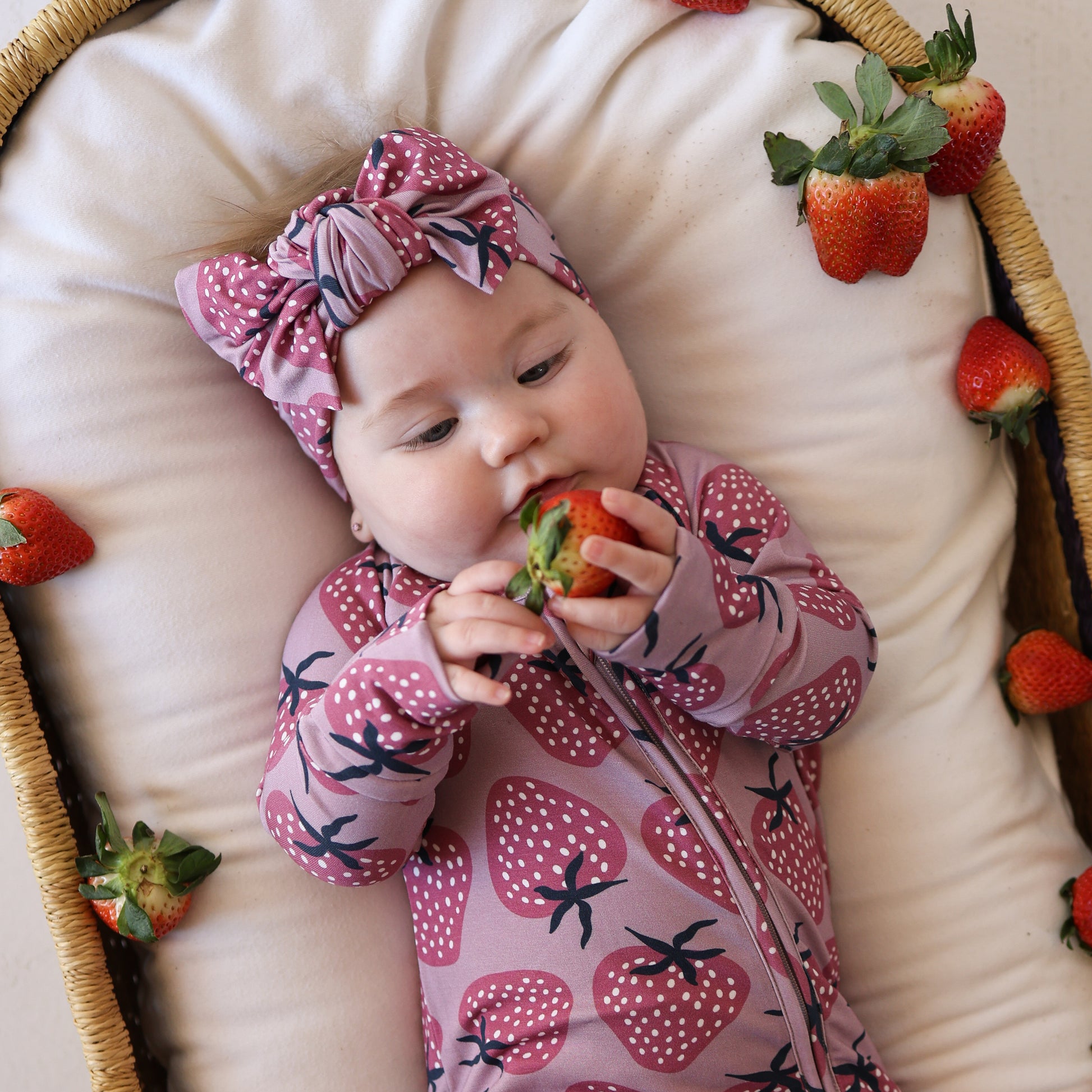 A baby lies in a basket, wearing forever french baby’s Bamboo Zip Pajamas | Matilda—a pink, hypoallergenic outfit with a strawberry pattern—and matching headband, surrounded by strawberries on a soft cushion.