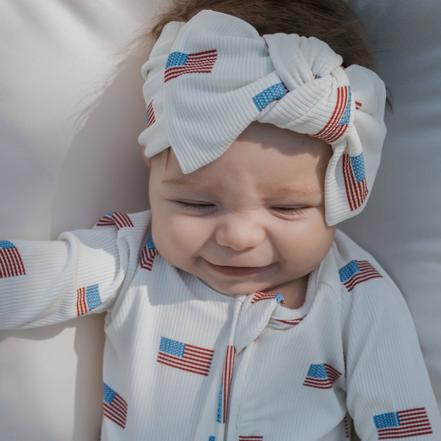 A baby in a white outfit and a forever french baby Bamboo Head Wrap | Americana Ribbed, decorated with American flag prints, smiles softly while lying down.