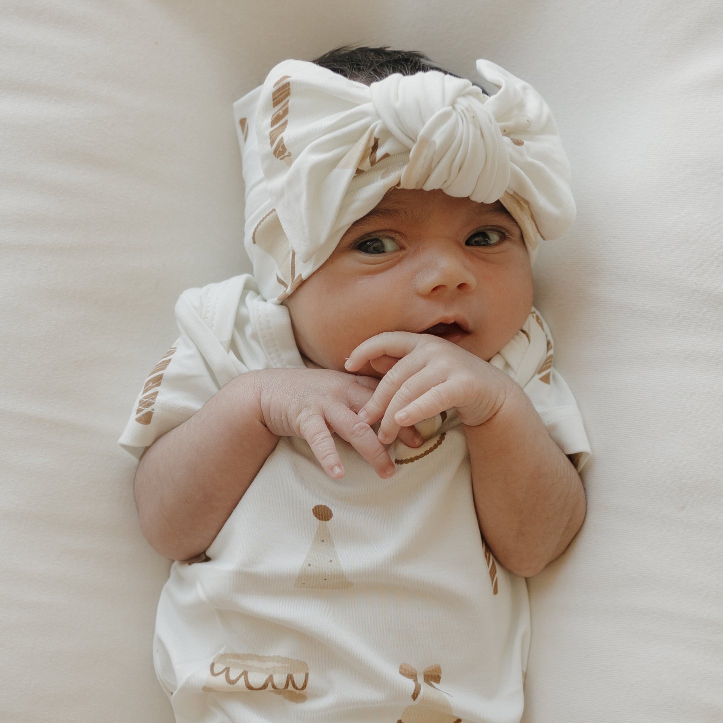 A newborn baby wearing a white headband with a large bow and the forever french baby Short Sleeve Bamboo Snapsuit | Birthday Girl lies on a white surface, hands near their mouth, gazing slightly to the side.