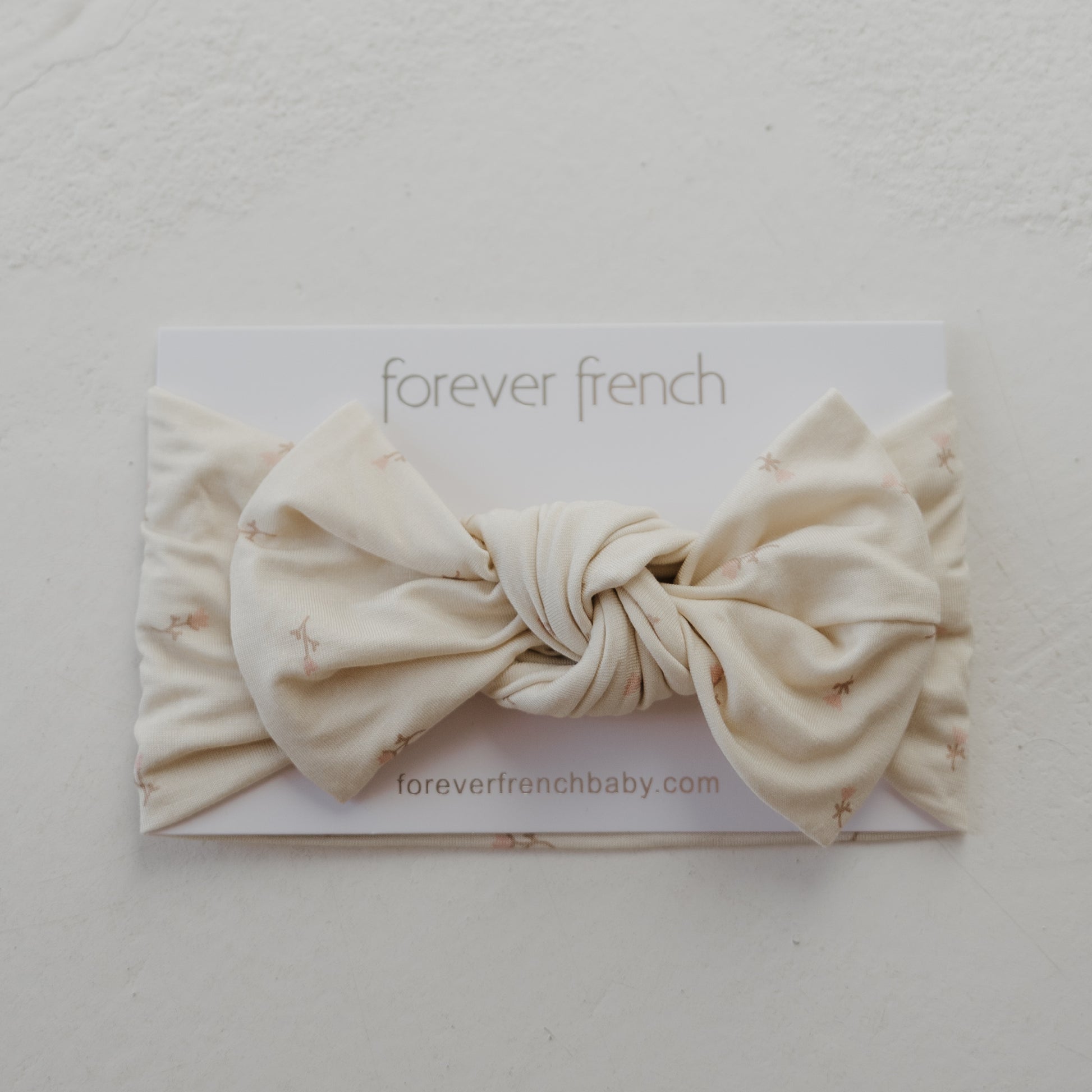 The Bamboo Head Wrap | Lucy by forever french baby is a cream-colored baby headband with a knotted bow and subtle star pattern, displayed on branded card. Visit foreverfrenchbaby.com.