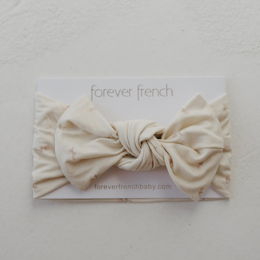 The Bamboo Head Wrap | Lucy by forever french baby is a cream-colored baby headband with a knotted bow and subtle star pattern, displayed on branded card. Visit foreverfrenchbaby.com.