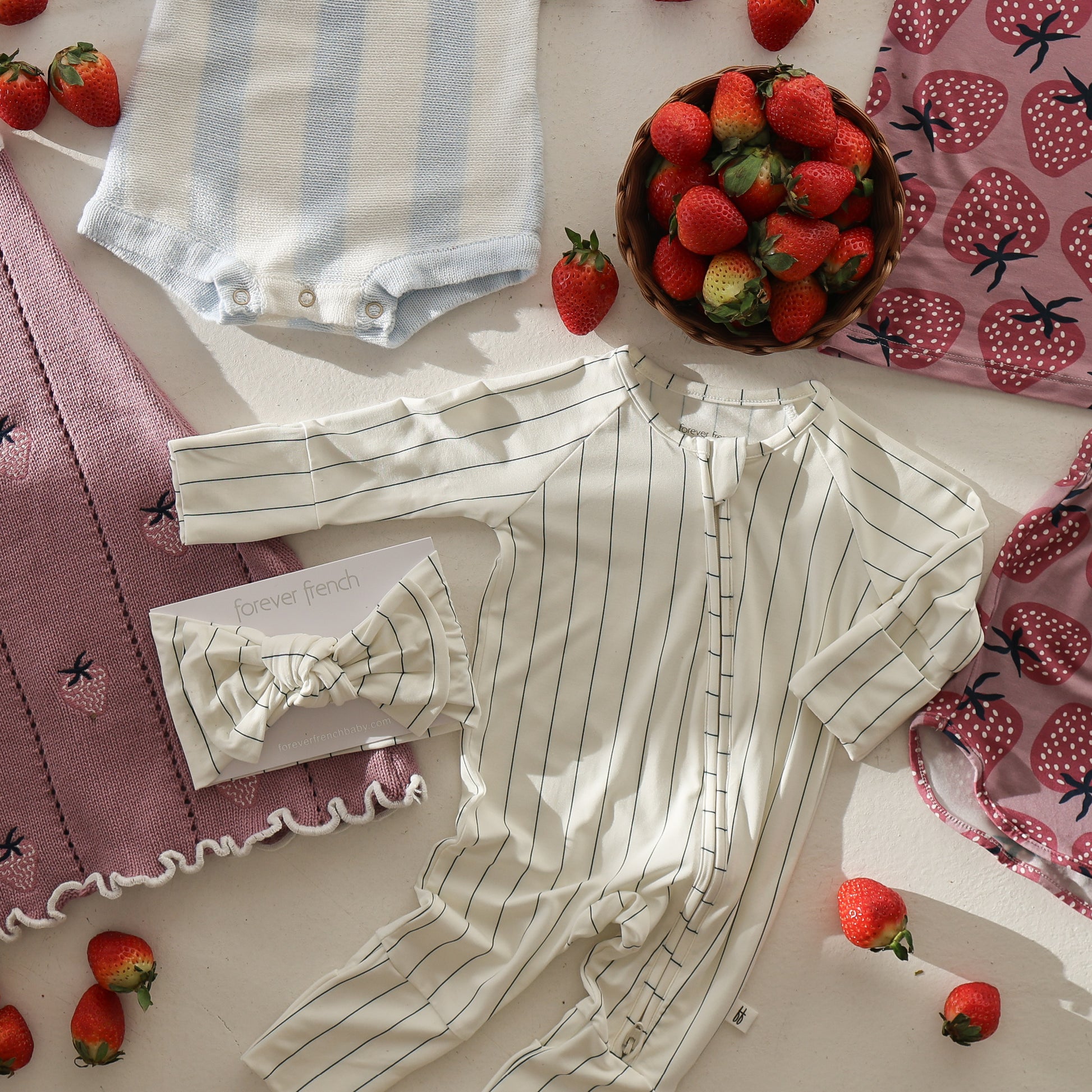 Flat lay of baby clothes and accessories, featuring the forever french baby Bamboo Head Wrap | Jack, a striped onesie, blue and white romper, strawberry-patterned pink blankets, a bowl of strawberries, and loose strawberries on white background.