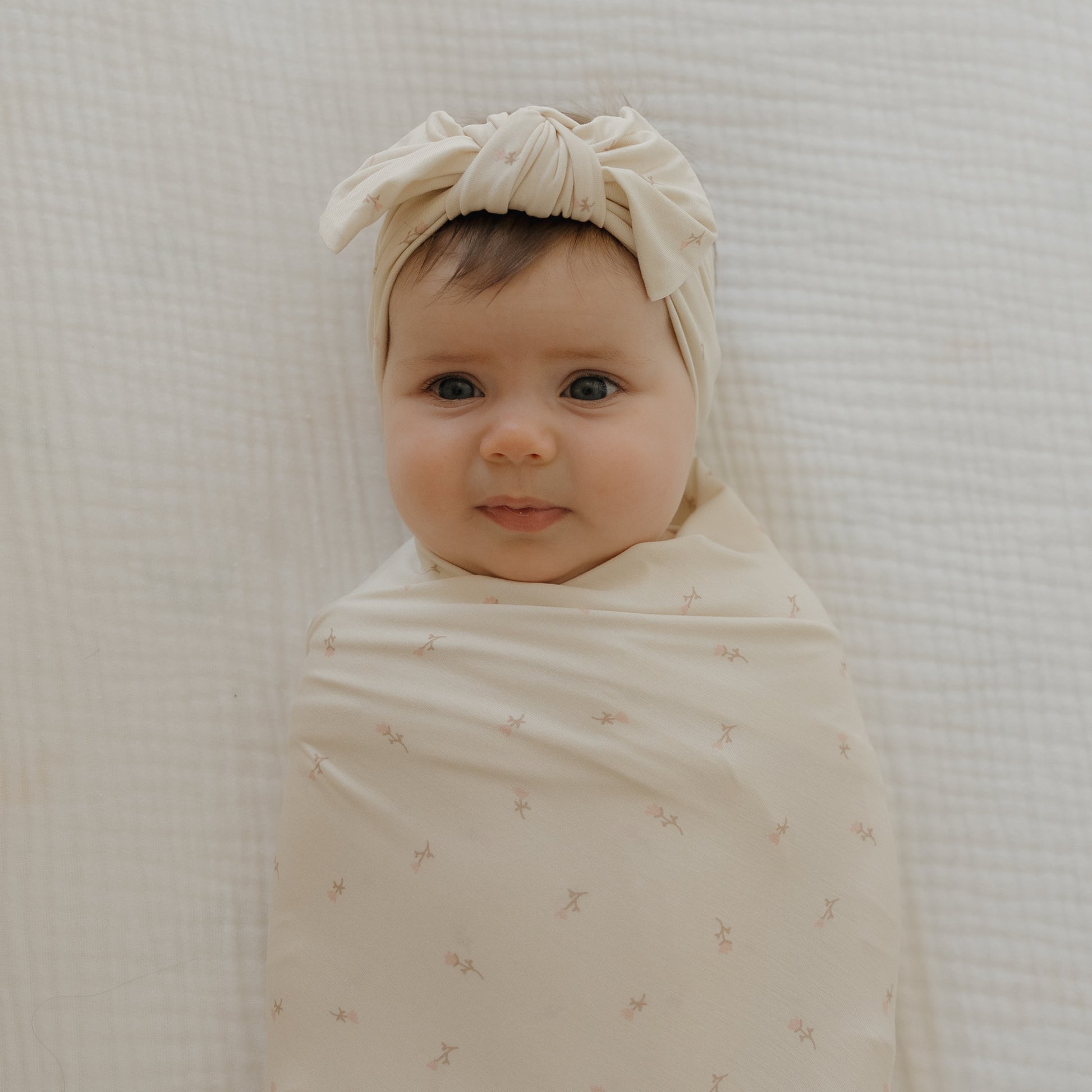 A baby with dark hair, wrapped in a cream swaddle and wearing the Bamboo Head Wrap | Lucy by forever french baby, lies on a textured white blanket, looking up with a gentle smile.