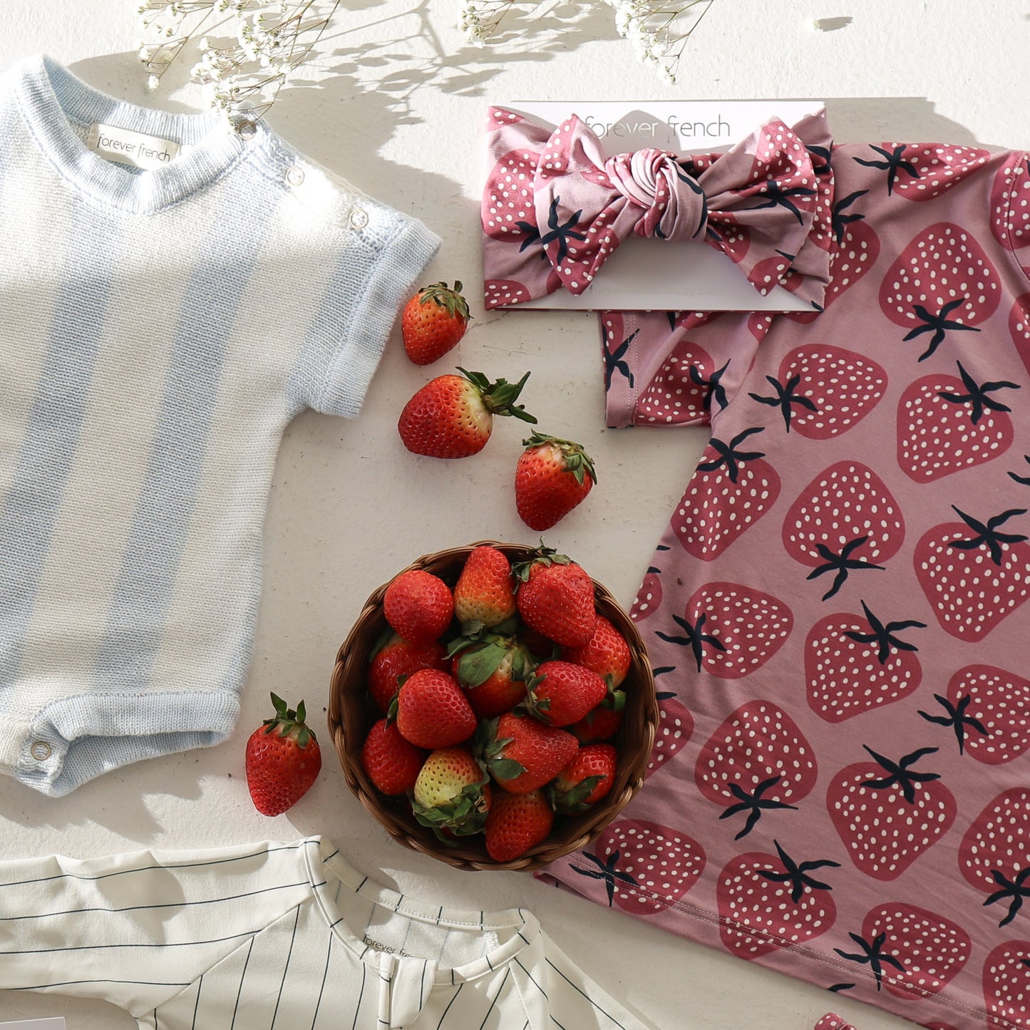 A flat lay of baby clothes and accessories, including a blue and white romper, a pink strawberry-patterned dress with the forever french baby Bamboo Head Wrap | Matilda, a striped outfit, and a bowl of strawberries on a white surface.