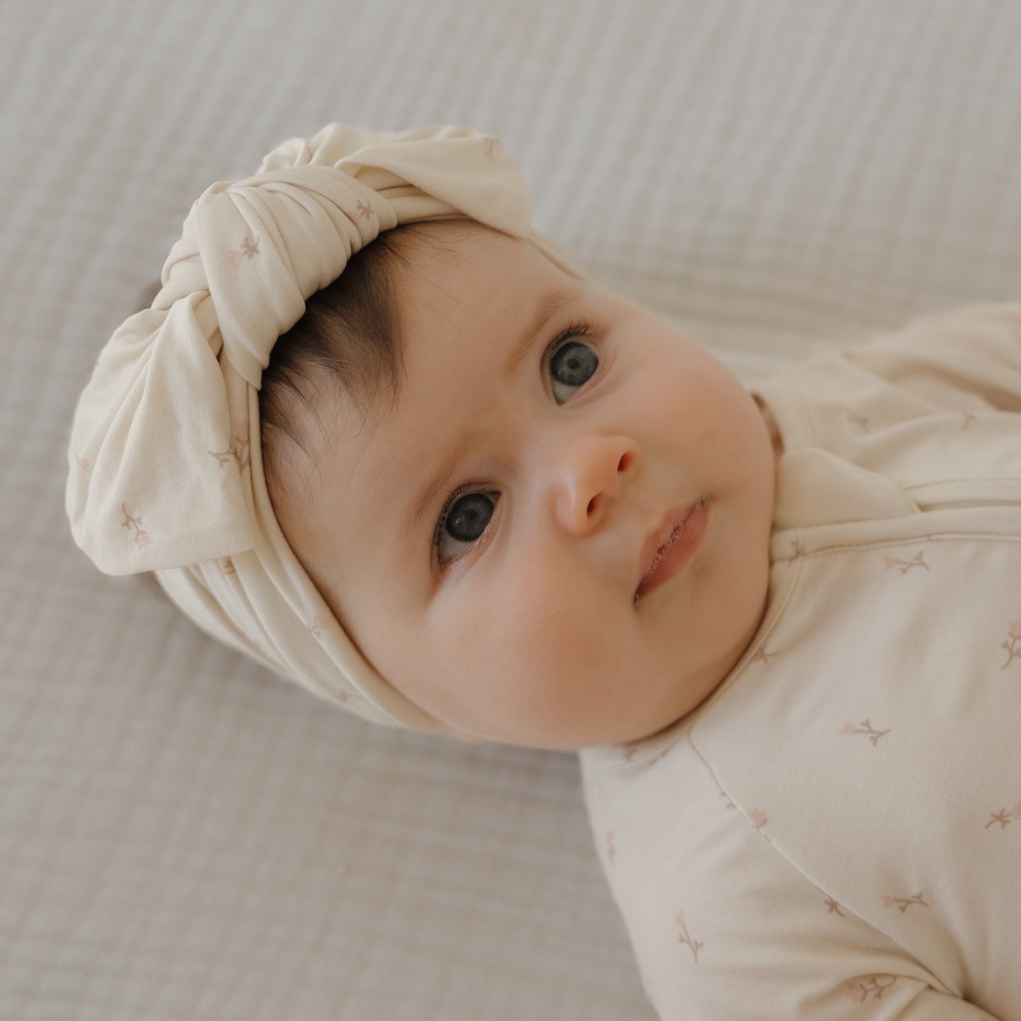 A baby with big blue eyes lies on a soft surface, wearing a cream outfit and the forever french baby Bamboo Head Wrap | Lucy, which features a large bow. The baby gazes upward with a calm expression.