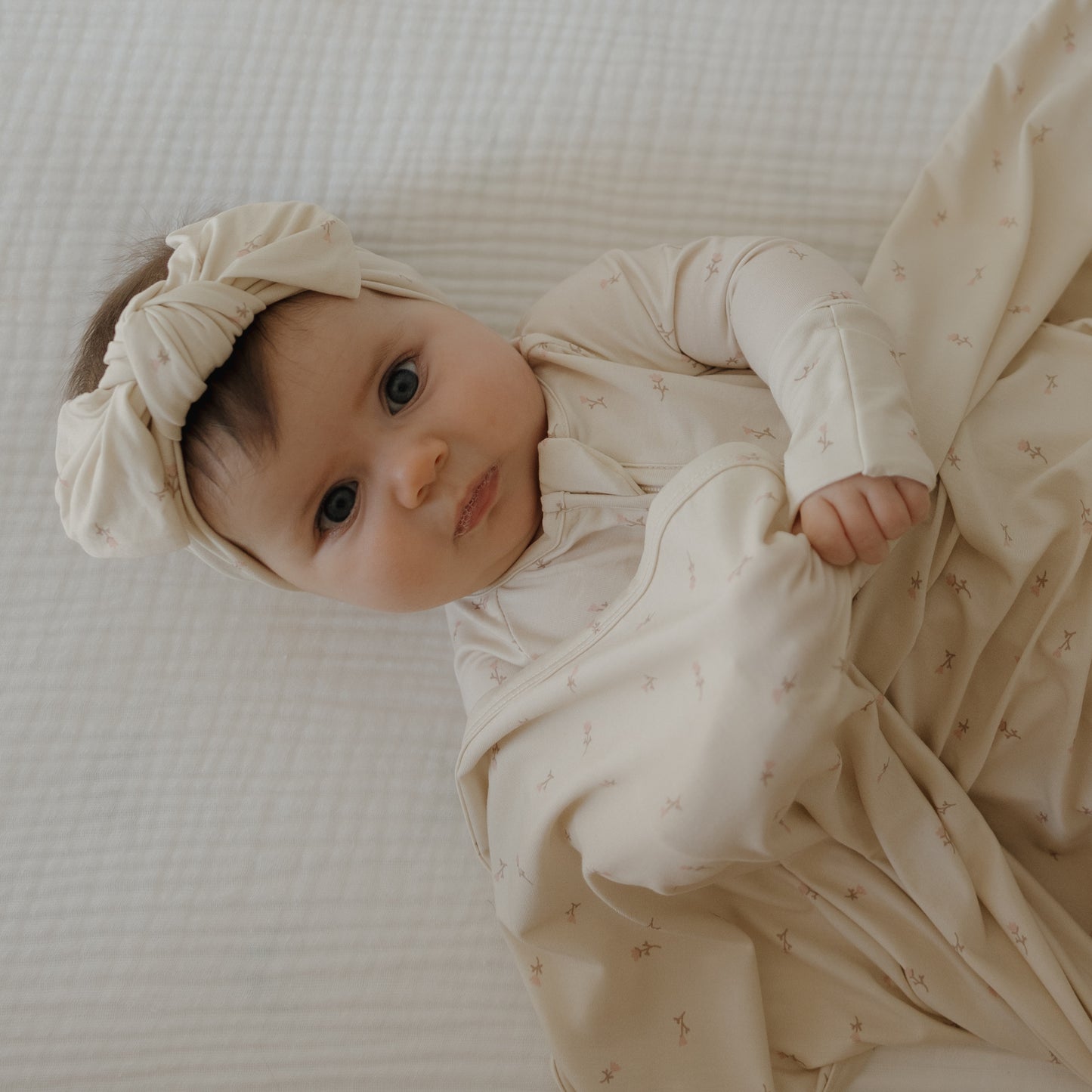 A baby in a cream outfit gazes upward while lying on a white textured blanket, wearing the forever french baby Bamboo Head Wrap | Lucy and grasping the soft fabric.