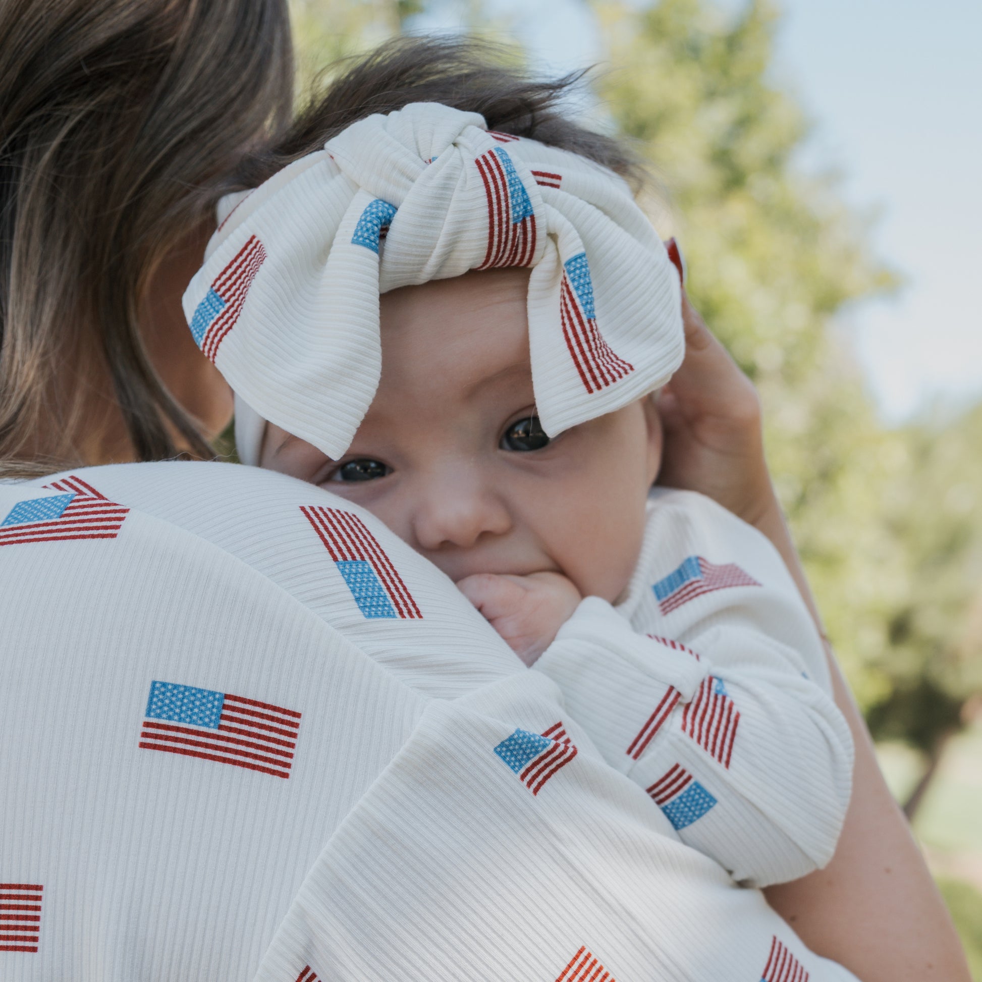 A baby in a forever french baby Bamboo Head Wrap | Americana Ribbed and American flag print outfit rests on a woman’s shoulder, both sporting matching flag patterns. Blurred trees and blue sky appear in the background.