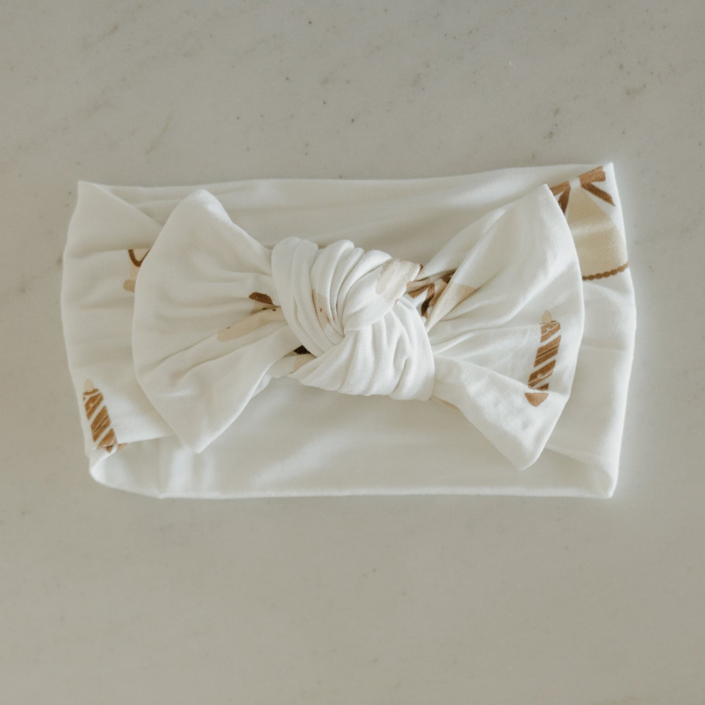 The Bamboo Head Wrap | Birthday Girl by forever french baby is a white fabric baby headband adorned with a large twisted bow in the center and subtle beige leaf patterns, elegantly displayed on a light surface—ideal as an elegant bamboo accessory or as the perfect birthday girl gift.