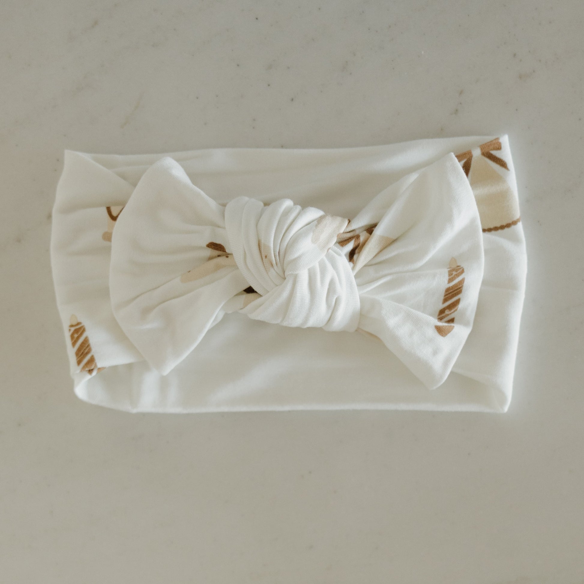 The Bamboo Head Wrap | Birthday Girl by forever french baby is a white fabric baby headband adorned with a large twisted bow in the center and subtle beige leaf patterns, elegantly displayed on a light surface—ideal as an elegant bamboo accessory or as the perfect birthday girl gift.
