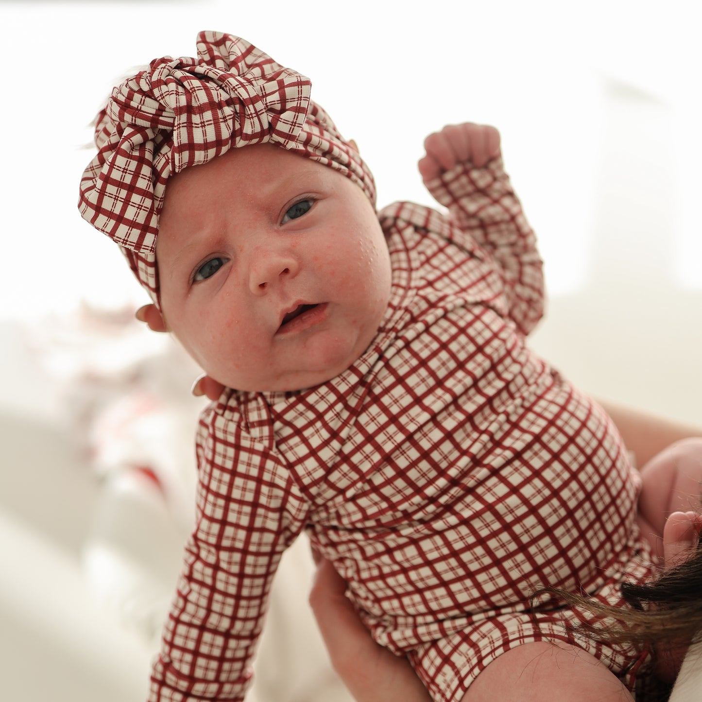 A baby wears the forever french baby Bamboo Head Wrap | Window Pane and a matching red and white plaid outfit, gazing at the camera with curiosity while being held against a softly blurred background.