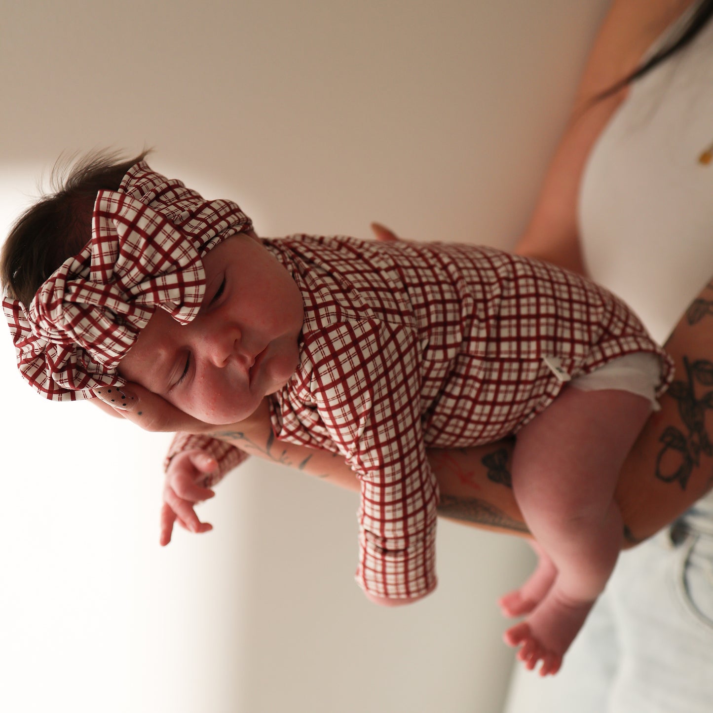 A baby in a matching red and white checkered outfit and a forever french baby Bamboo Head Wrap | Window Pane with an adjustable bow sleeps peacefully, cradled in tattooed adult arms against a plain background.