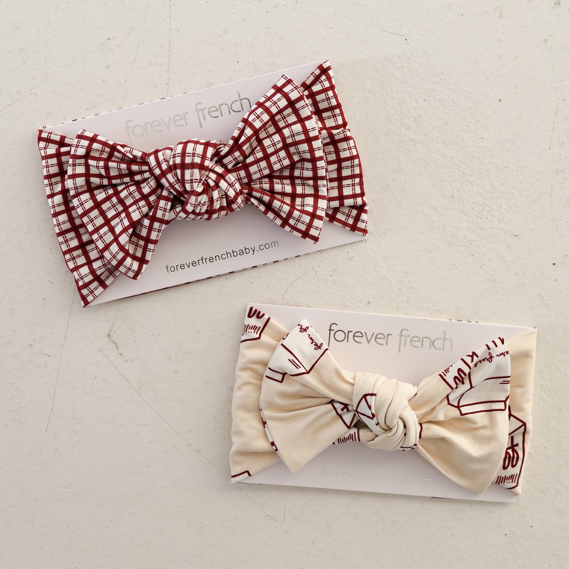 Two forever french baby Bamboo Head Wraps on branded cards—one features a red and white plaid adjustable bow ("Window Pane"), the other is cream with maroon abstract patterns. Both soft wraps are displayed on a light, textured surface.