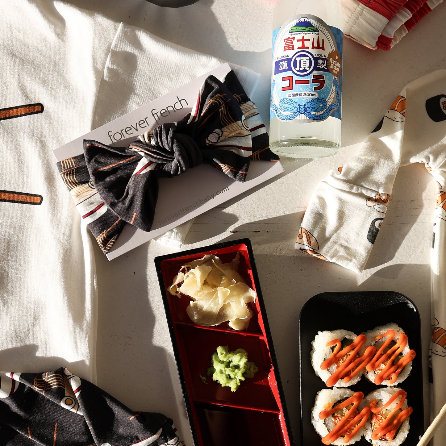 A flat lay featuring sushi rolls with orange sauce, ginger and wasabi tray, a black patterned bow tie, Japanese soda, fabric with illustrations, and the forever french baby Bamboo Head Wrap | Ramen—all in sunlight.