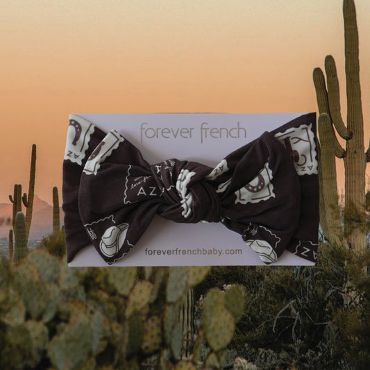 The Bamboo Head Wrap | With Love from AZ by forever french baby, an adjustable dark brown wrap with white Arizona-inspired prints, is displayed on branded packaging against a sunset desert scene with cacti.