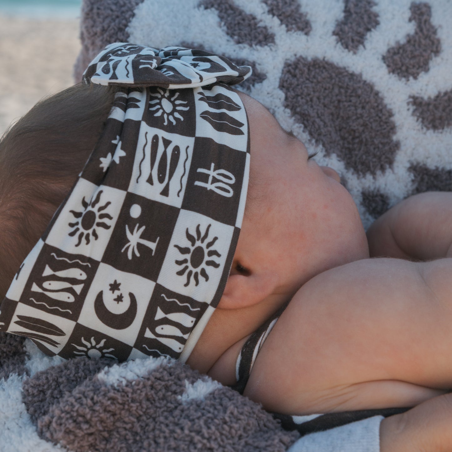 A baby in the forever french baby Bamboo Head Wrap | Summer Check, featuring playful patterns, lies peacefully on a matching towel.