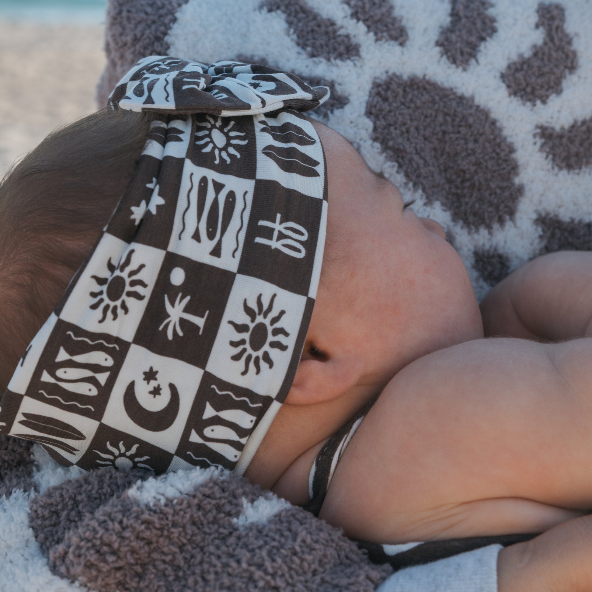 A baby in the forever french baby Bamboo Head Wrap | Summer Check, featuring playful patterns, lies peacefully on a matching towel.