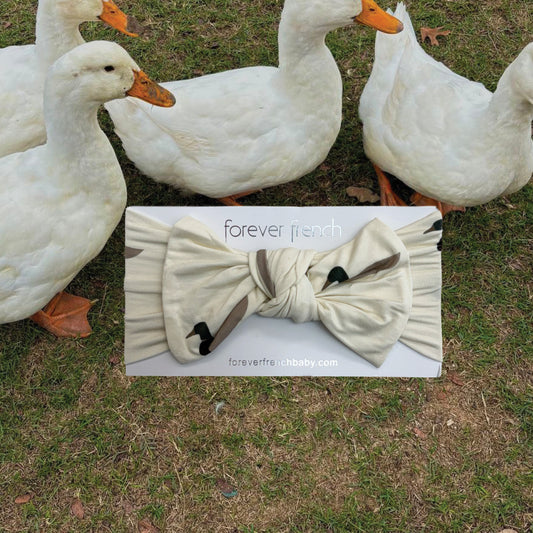 Four white ducks stand on grass behind a forever french baby Bamboo Head Wrap | Mallard, adorned with a bow and placed on the ground in front of them. This charming head wrap showcases a stylish mallard-inspired white and black pattern.