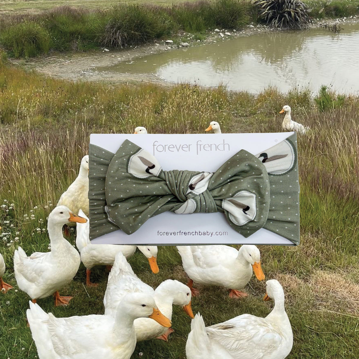 A forever french baby Bamboo Head Wrap | Mallard Frames is displayed in front of a pond, with white ducks standing on the grass. The charming bamboo head wrap features a playful duck print.
