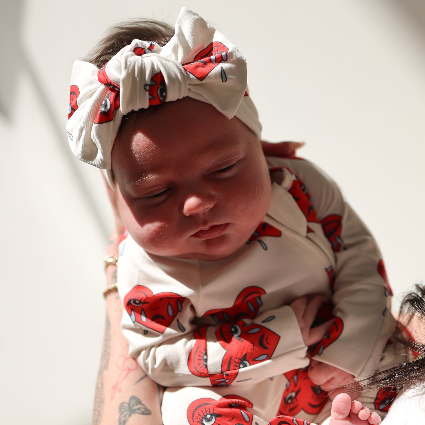 A baby in a cream outfit and the forever french baby Bamboo Head Wrap | Sad Girl, featuring red cartoon eye designs, is gently held by an adult as sunlight softly casts gentle shadows over them.