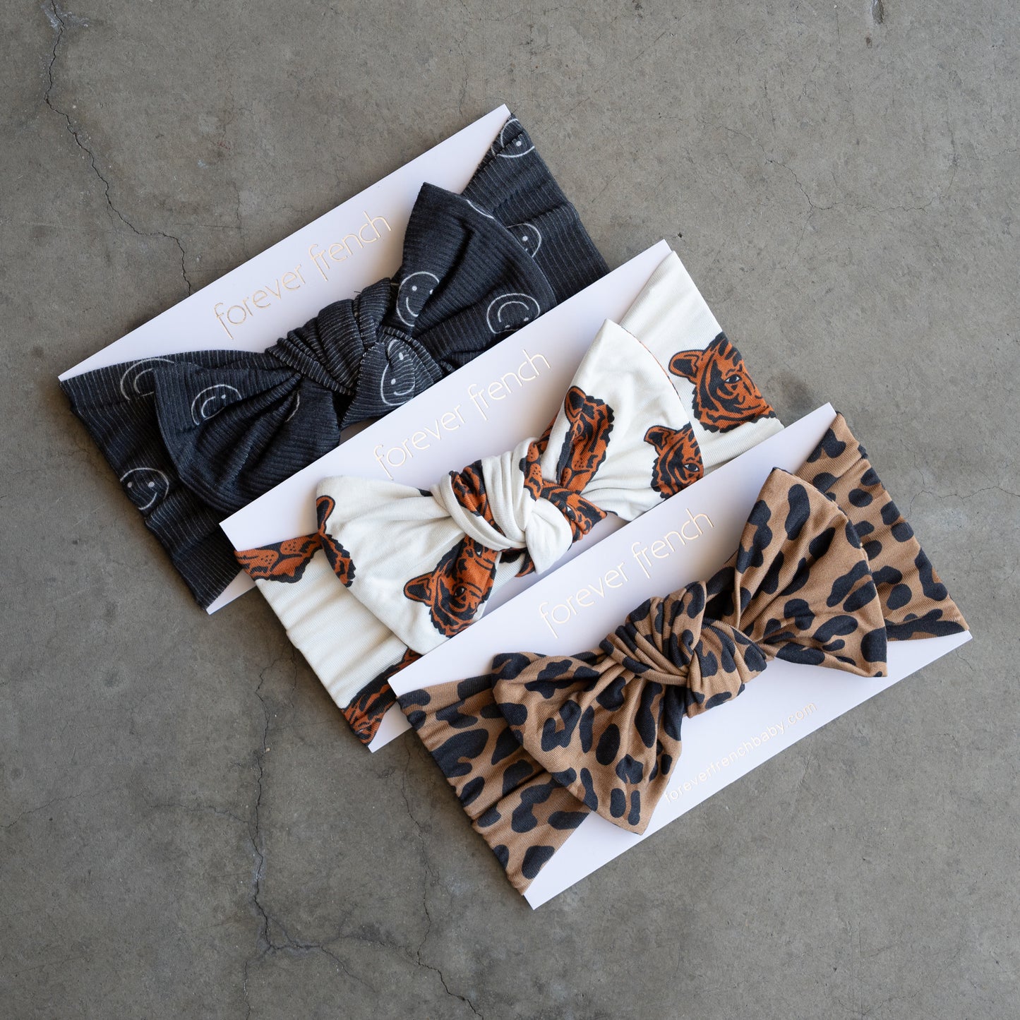 The Bamboo Head Wrap | Tigers by forever french baby features knotted, adjustable headbands in black moons, white tigers, and brown leopard spots, displayed on branded cards atop a concrete surface.